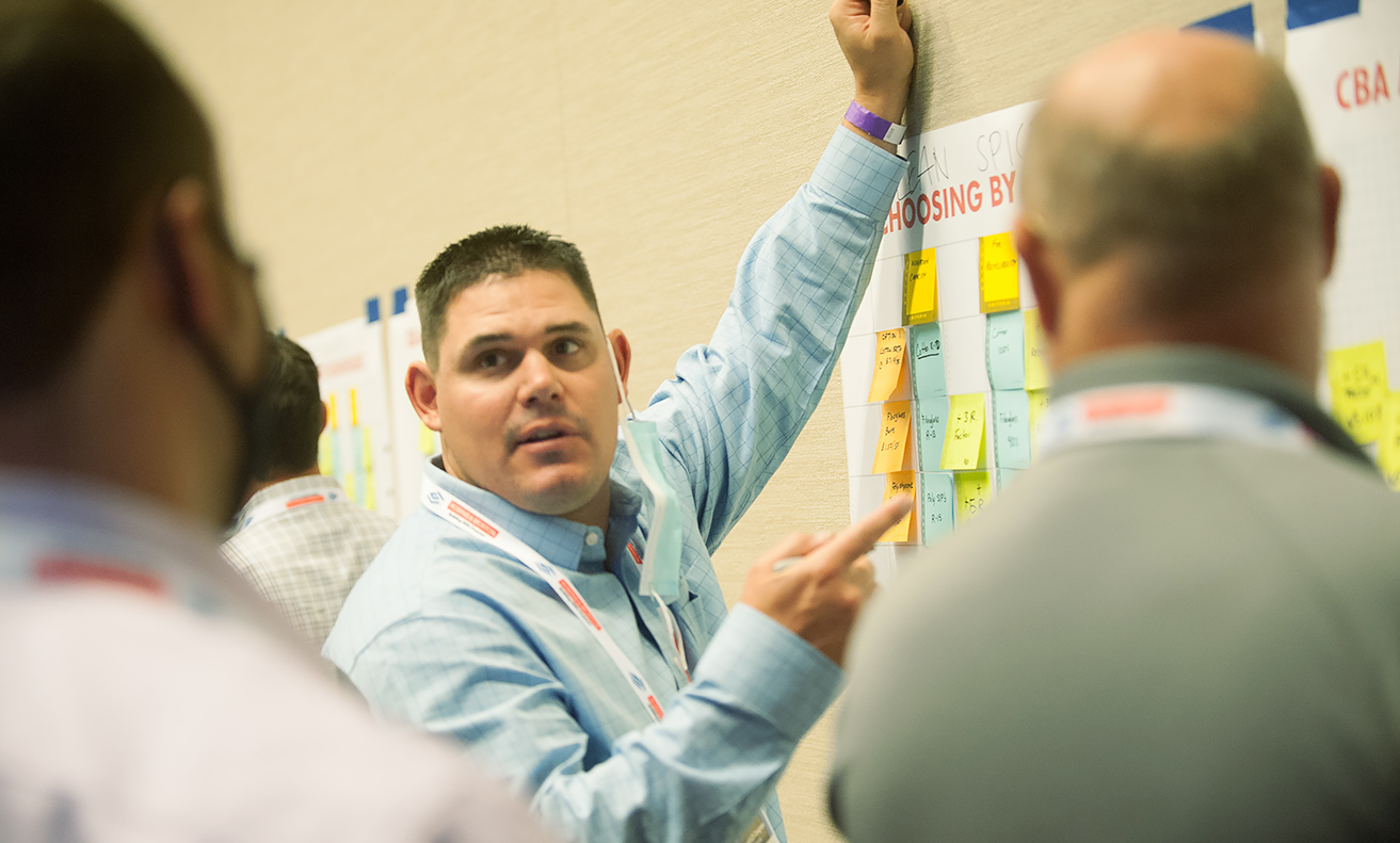man in light blue shirt using both arms to point at a wall with sticky notes on it
