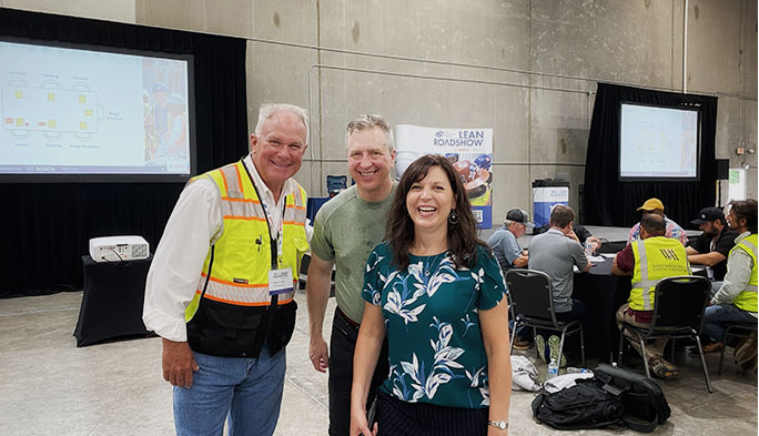 Two men and a woman smile for the camera in front of the Lean Roadshow poster.