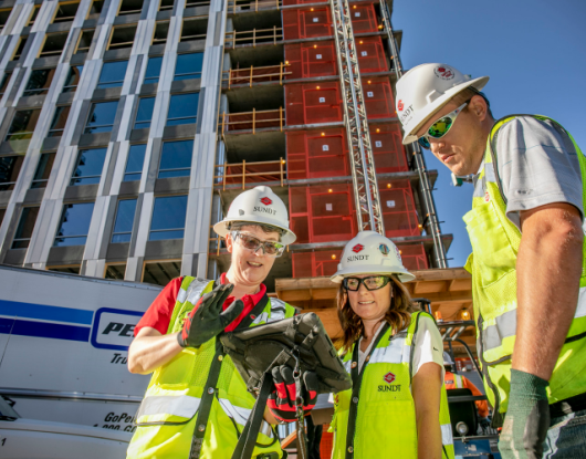 Two women and a man in yellow vests, white hard hats, and safety glasses look at an iPad stating their Lean mission.