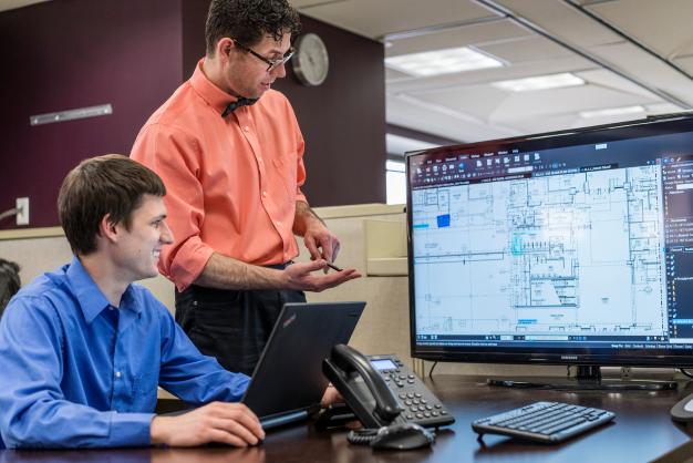 A man in a red button-down stands over a man sitting at a desk in a blue button-down while he does work on a computer.